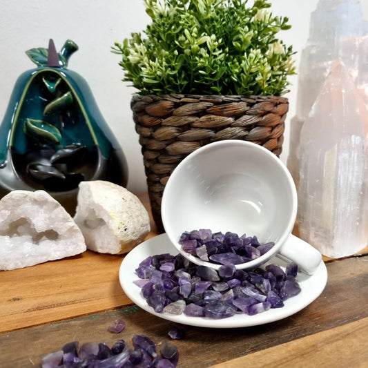 A white teacup spills small, tumbled amethyst stones onto a white saucer on a wooden surface. Background elements include an incense holder, crystals, and a potted plant.