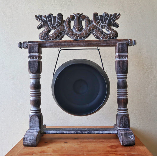 A black gong suspended in an ornately carved wooden stand resembling intertwined dragons, situated on a wooden table against a plain beige wall.