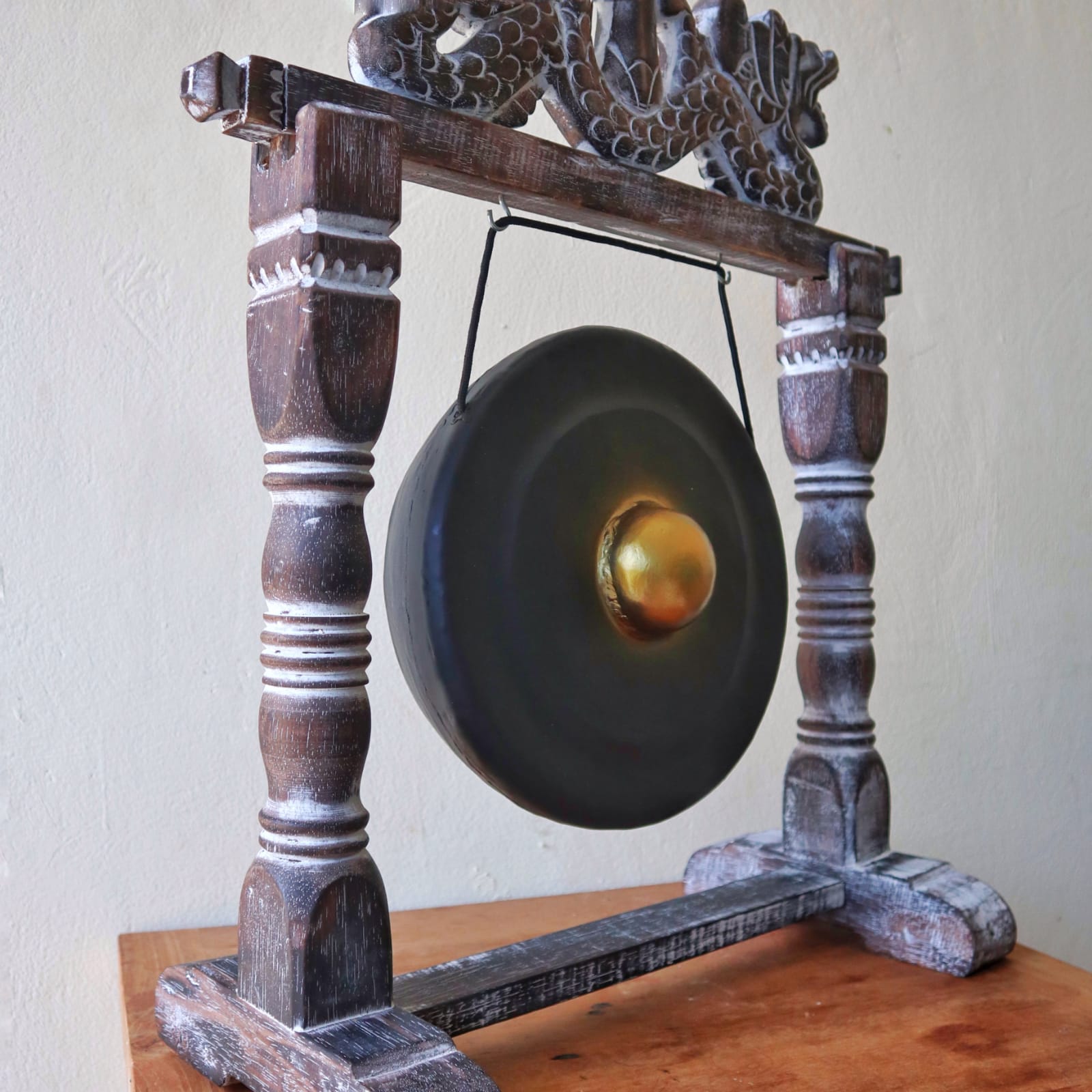 A decorative gong hangs from an ornate wooden frame with carved patterns, resting on a wooden table against a plain light-colored wall.