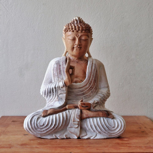 A wooden Buddha statue sits cross-legged on a wooden surface, with one hand raised in a gesture, against a plain textured gray background.