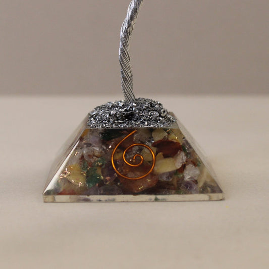 A pyramid, made of resin filled with colorful stones and a copper swirl, supports a small metal tree. It sits on a light surface against a light background.