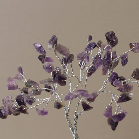 A tree sculpture stands with silver wire branches adorned with small, irregularly shaped, purple amethyst stones, all against a tan background.