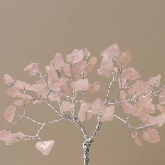 A wire tree displays pink, gemstone "leaves" on thin, silver branches. The tree is set against a solid, light tan background.