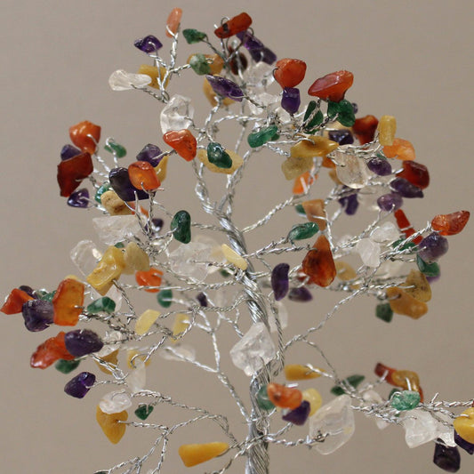 A wire tree displays colorful, polished stones on its branches, simulating leaves. The tree stands against a neutral, slightly reflective backdrop.