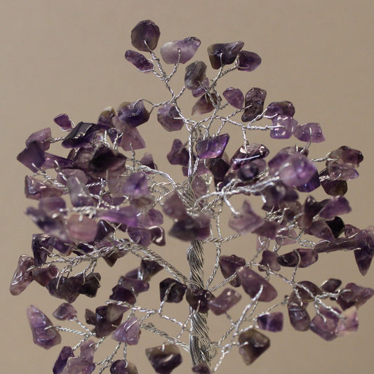A decorative tree with silver wire branches is adorned with polished amethyst stones. The tree stands against a plain, light brown background.