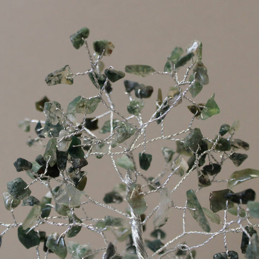 A silver wire tree displays green stones as leaves, with the tree’s trunk positioned at the bottom against a beige background.