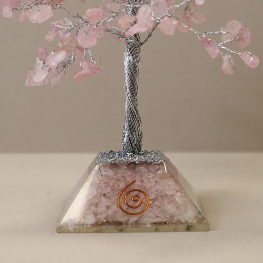 A decorative tree with a silver wire trunk and rose quartz "leaves" stands on a pyramid-shaped base containing quartz pieces and a copper spiral. The tree sits on a light surface against a neutral background.