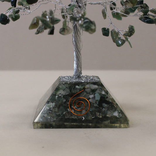 A decorative gemstone tree stands on a light surface. The tree has green gemstone leaves and a twisted wire trunk rising from a pyramid-shaped, green stone base with a copper spiral accent.