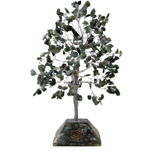 A gemstone tree stands on a faceted gemstone base. The tree's trunk and branches are silver wire, with irregular green gemstones as leaves against a white background.
