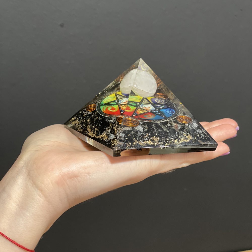 A black, glittery pyramid with a colorful symbol and quartz point rests in a fair-skinned hand. The background is a dark gray.