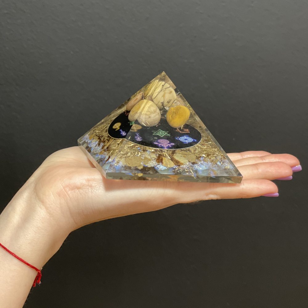 A pyramid of mixed materials, including rocks and metal flakes, rests in a person's hand, against a dark background. A black oval with colorful symbols adorns the inner part.