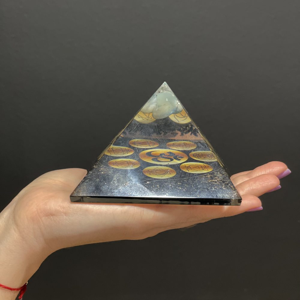 A pyramid-shaped orgonite, embedded with symbols, is held in a palm against a dark background.