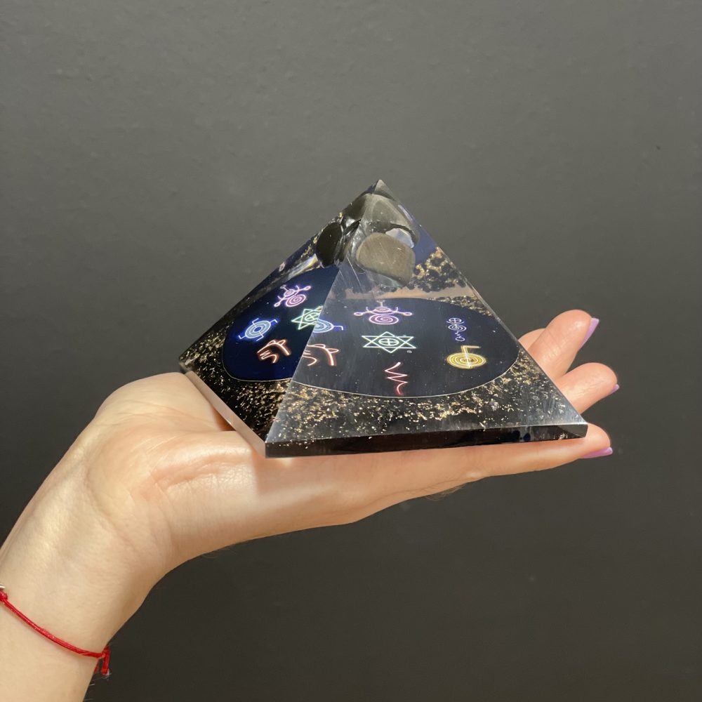 A black orgonite pyramid with stones and colorful symbols is being held in a hand against a dark gray backdrop.