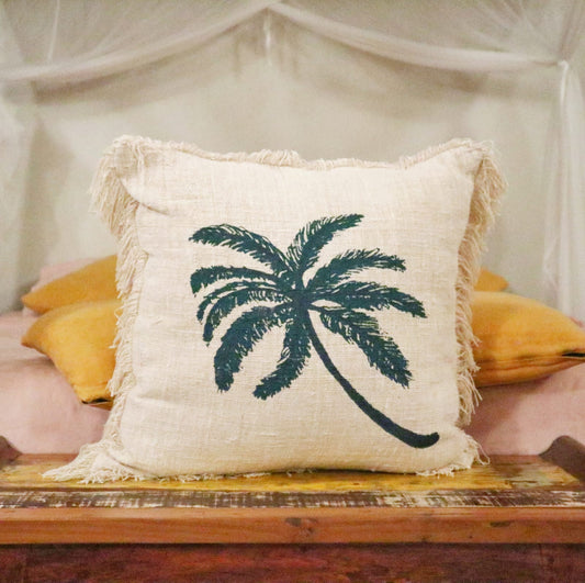 A beige pillow, featuring a dark-green palm tree print, rests on a wooden surface. It is situated on a bed with mustard-yellow pillows.