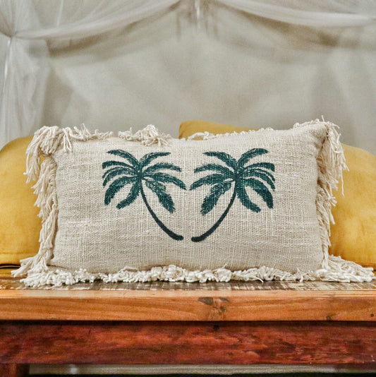 A beige pillow, decorated with two dark-green palm trees, rests on a wooden bench. The bench is situated in front of a sheer, light-beige curtain.
