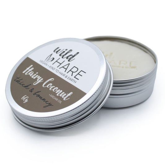 Aluminum tin of hair balm with lid partially removed revealing solid balm, lid resting at an angle on a plain white background. Text: wild HARE VEGAN + KIND TO HAIR & EARTH Hairy coconut thick & bouncy +ARGAN OIL 60g