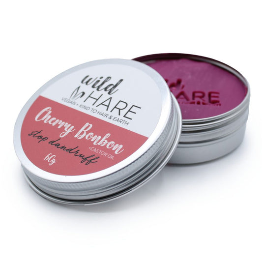 A small round metal tin of hair wax sits with its lid partially removed to reveal magenta wax on a white surface; label reads "wild HARE VEGAN + KIND TO HAIR & EARTH Cherry Bonbon Stop dandruff +CASTOR OIL 60g".