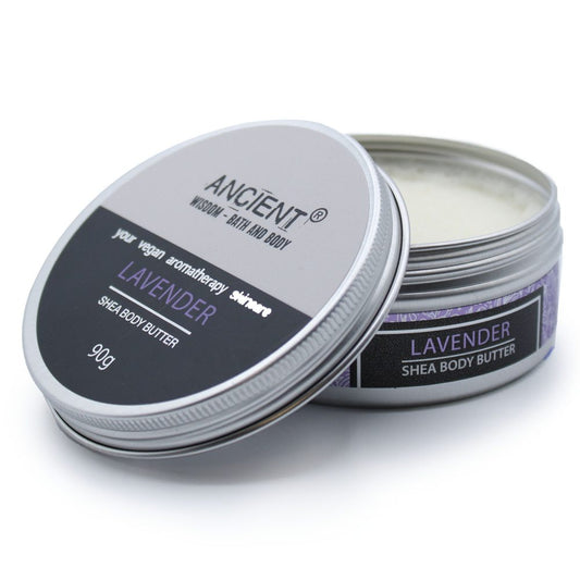 A round metal tin of lavender shea body butter with its lid partially removed revealing cream, resting on a white surface; text: "ANCIENT® WISDOM - BATH AND BODY", "your vegan aromatherapy skincare", "LAVENDER", "SHEA BODY BUTTER", "90g", side label "LAVENDER SHEA BODY BUTTER".