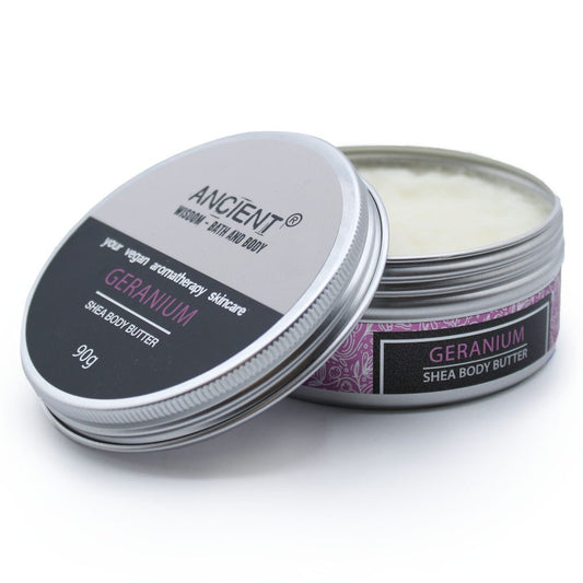 A metal tin of shea body butter sits partially open revealing cream on a white surface, label reads ANCIENT® WISDOM - BATH AND BODY your vegan aromatherapy skincare GERANIUM SHEA BODY BUTTER 90g