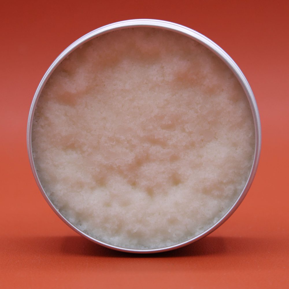 An open, silver-colored tin holds a granular, white balm filling the container. The tin sits on a smooth, solid orange surface.