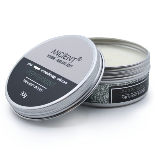 A 90g tin of "ANCIENT WISDOM - BATH AND BODY" peppermint shea body butter has its lid askew to reveal a white cream inside. Text on the lid reads, "your vegan aromatherapy skincare PEPPERMINT SHEA BODY BUTTER." Another sticker on the side of the pot also reads, "PEPPERMINT SHEA BODY BUTTER"