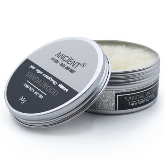 Open tin of Sandalwood Shea Body Butter sits on a white surface. The tin lid reads "ANCIENT. WISDOM-BATH AND BODY. Your vegan aromatherapy skincare. Sandalwood. Shea Body Butter. 90g." The tin's label reads "Sandalwood Shea Body Butter."