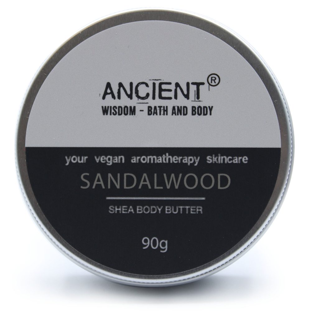 A round tin of body butter sits on a white surface, labeled "ANCIENT, WISDOM - BATH AND BODY, your vegan aromatherapy skincare, SANDALWOOD, SHEA BODY BUTTER, 90g."