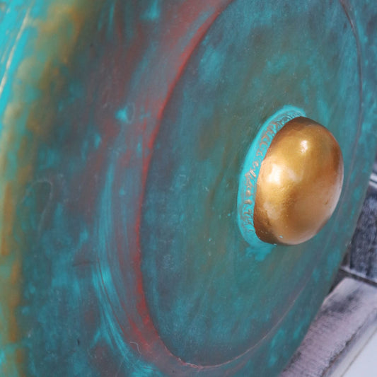 A large, round, turquoise shield with a central gold dome is mounted against a patterned surface, displaying a textured, metallic finish in a close-up view.