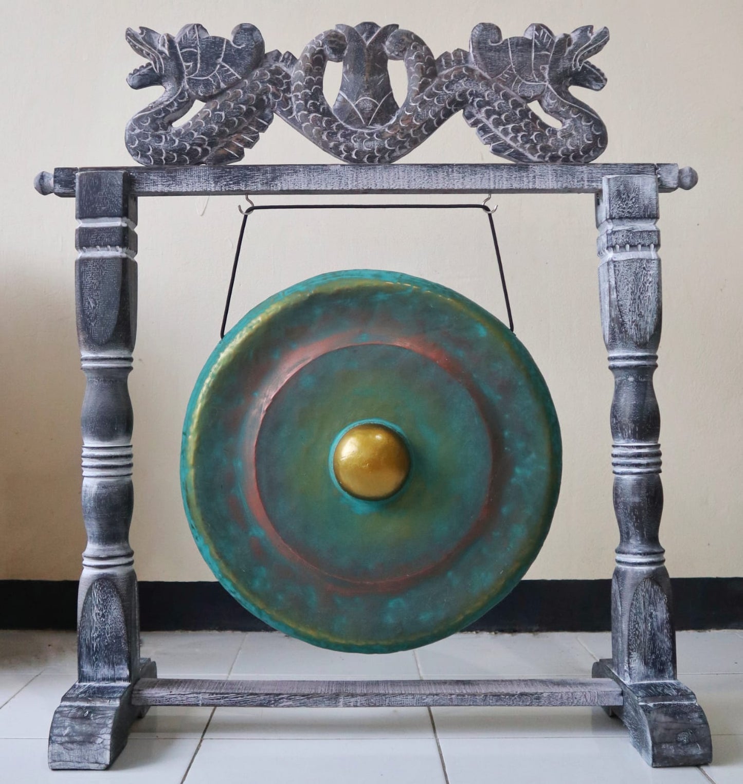 A decorative gong with a greenish surface and golden center rests on a wooden stand, adorned with carved dragon motifs, in a tiled indoor setting.