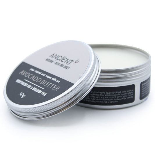 Metal tin of avocado butter sits with lid leaning open on a white surface; label reads "ANCIENT® HAND · BATH AND BODY" "your natural and vegan skincare" "AVOCADO BUTTER" "MOISTURIZES DRY & DAMAGED SKIN" "90g".