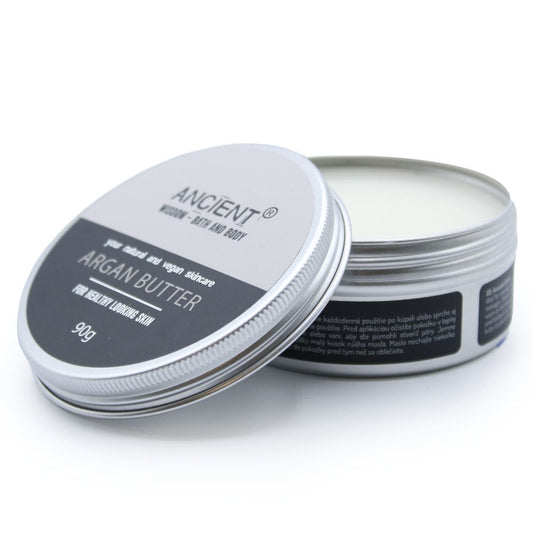 Round metal tin of Argan Butter sits open showing cream inside on a white background; lid leans against tin; text on lid reads "ANCIENT®" "pure natural and vegan skincare" "ARGAN BUTTER" "FOR HEALTHY LOOKING SKIN" "90g" and small side label text is too small to read.