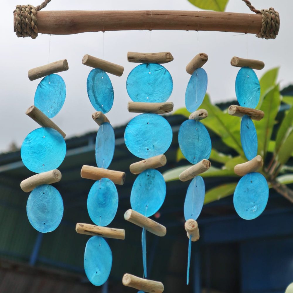 A wind chime hangs from a wooden bar; light blue discs and small wooden pieces are strung, creating a mobile effect; the setting is outdoors with greenery in the background.
