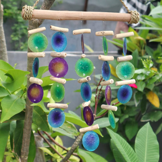 A mobile of iridescent, circular shell pieces hangs from driftwood, strung in vertical rows. It's suspended outdoors amongst lush green foliage.
