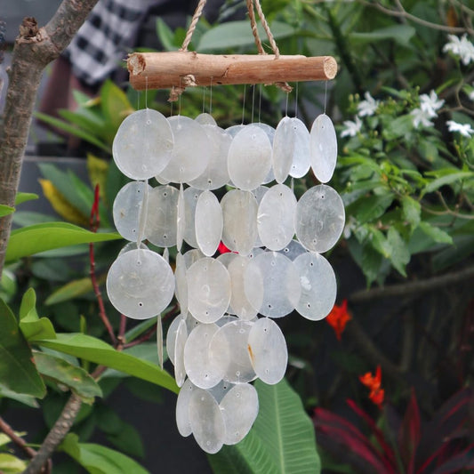 A shell wind chime hangs from a wooden branch, gently swaying amidst lush green foliage and small white and red flowers.