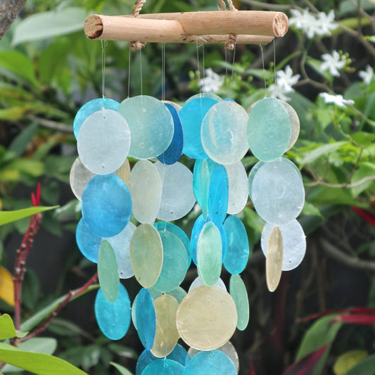 A wooden-framed wind chime, comprised of numerous circular shell pieces in varying shades of blue and beige, hangs gently amidst lush green foliage.