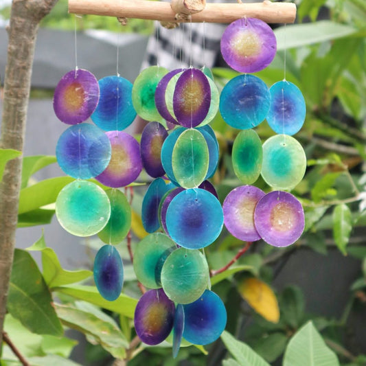 A mobile of circular, iridescent ornaments hangs from a wooden rod. The ornaments are various shades of purple, teal, and green. The mobile is suspended amidst lush green foliage.
