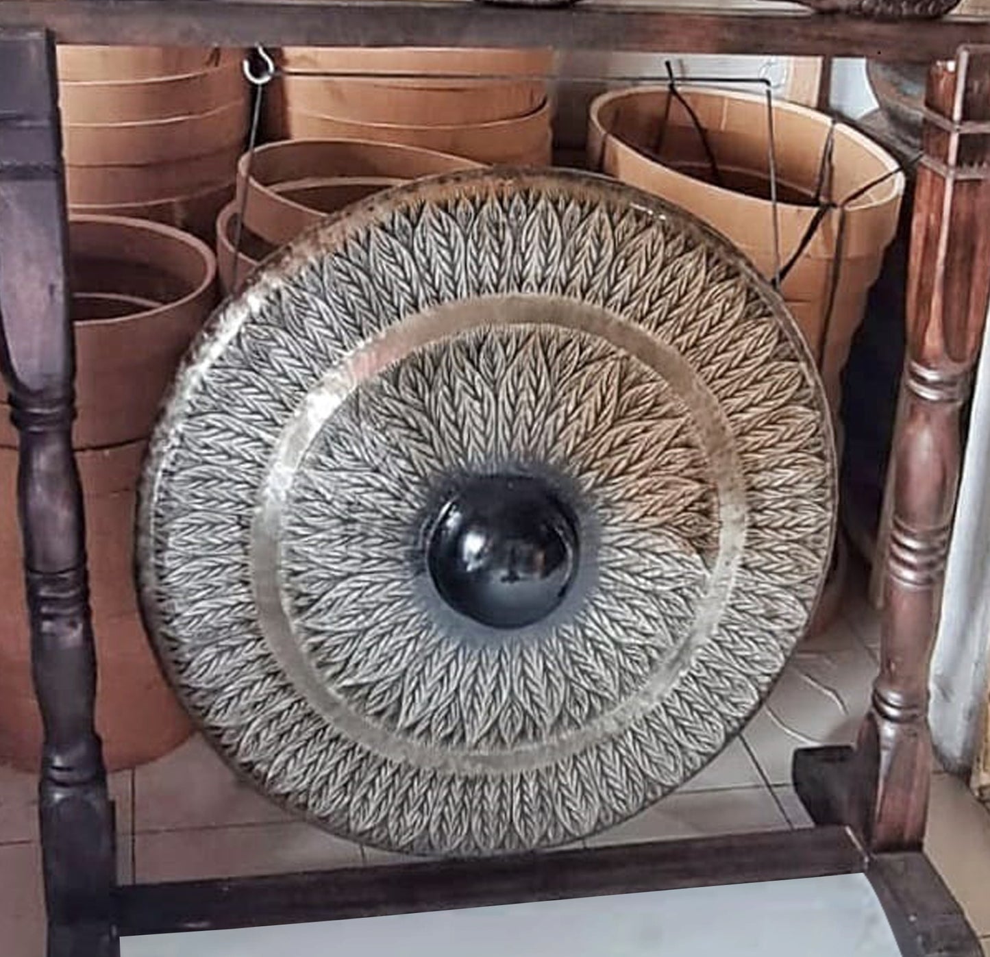 A decorative gong with intricate leaf patterns hangs in a wooden frame, surrounded by several stacked tan sand sieves, set on a tiled floor.