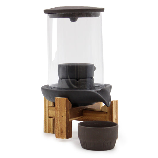 A glass bird feeder sits atop a wooden stand, featuring a dark roof and spout for water collection, with a matching small dish placed in front.