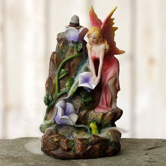 A fairy figurine, dressed in a pink gown, leans over purple flowers on rocky terrain. A small waterfall flows beside her, creating misty smoke in a soft, blurred background.