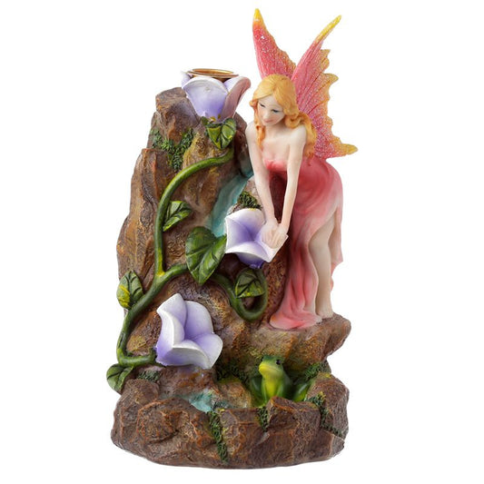 A figurine of a fairy in a pink dress kneels atop a rocky structure with green leaves and purple flowers. A small frog sits near the base, creating a whimsical scene.