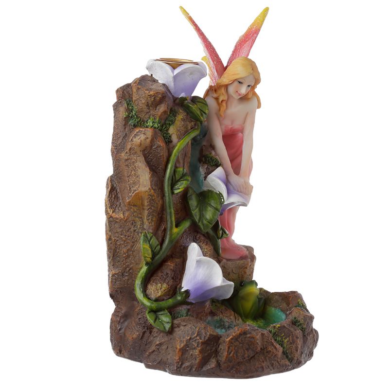 A fairy figurine with colorful wings sits on a rocky base, surrounded by blooming flowers and green leaves, creating a whimsical, natural scene.