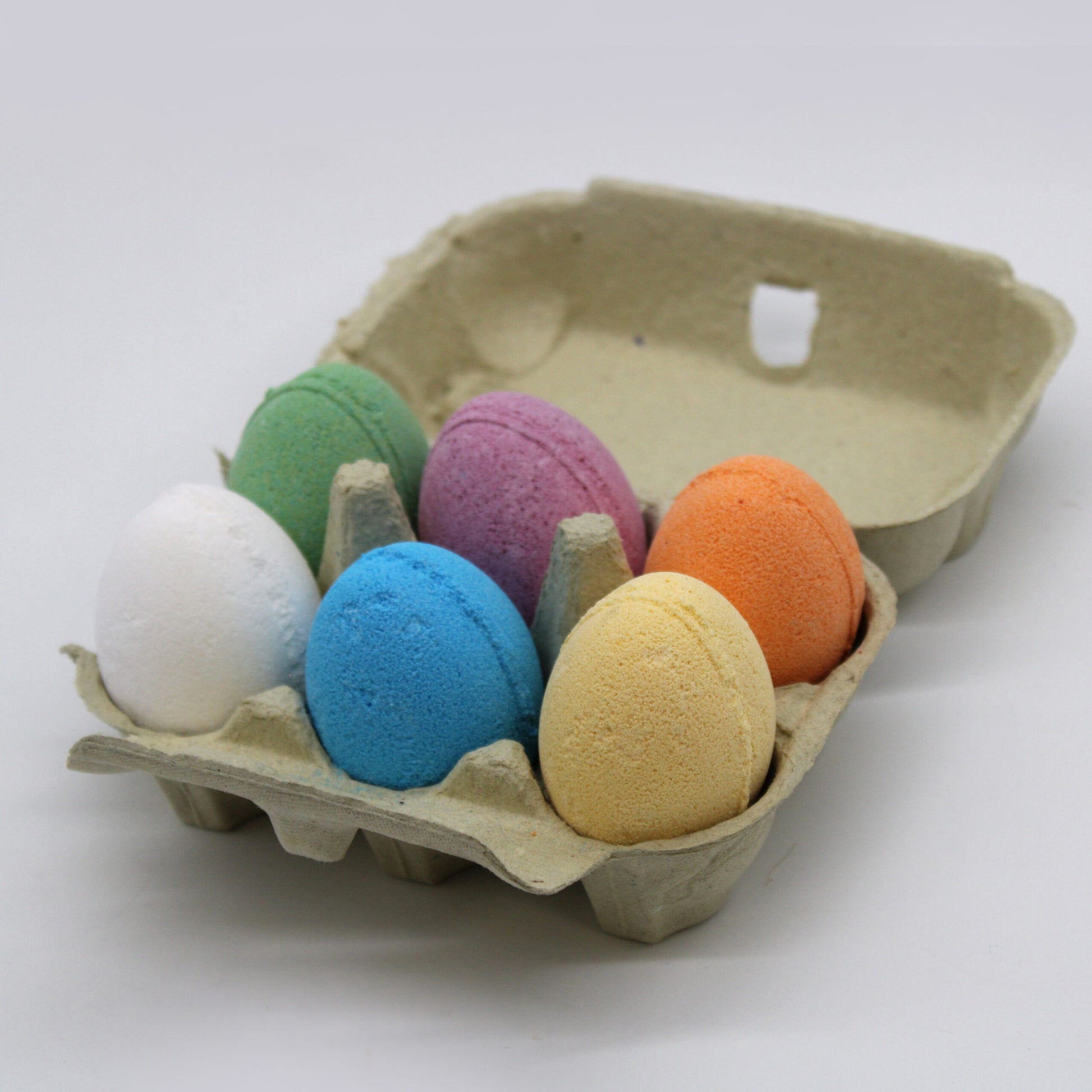A carton holds six colorful oval objects resembling eggs—white, green, blue, purple, orange, and yellow—resting in an open egg carton against a plain, light gray background.