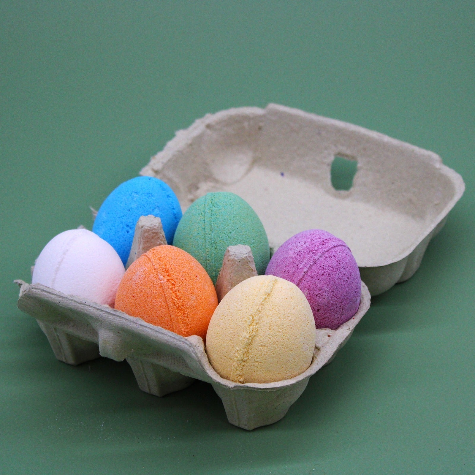 Colorful bath bombs are neatly arranged in an open cardboard egg carton on a plain green background.