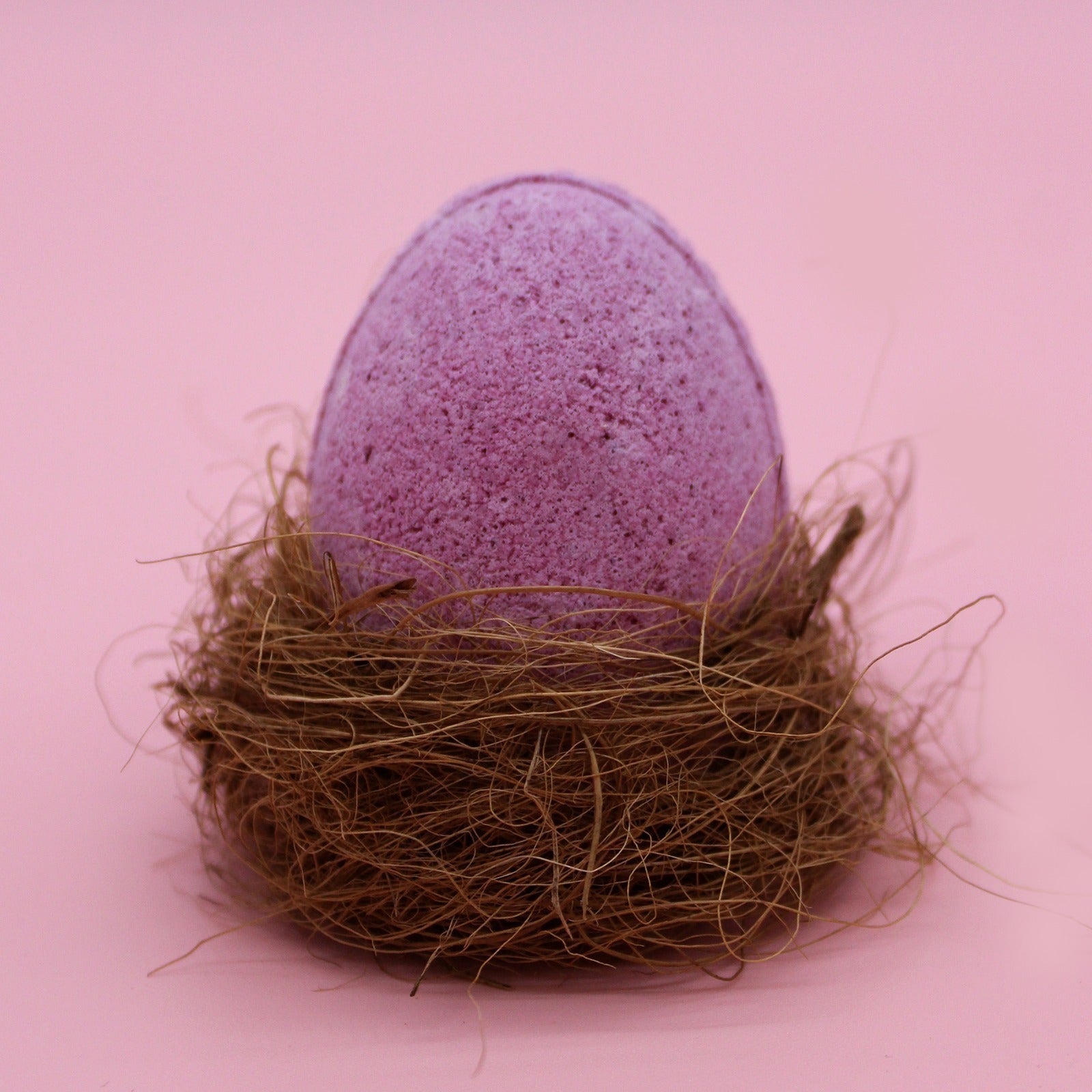 A pink, egg-shaped bath bomb rests in a small nest of straw-like material, set against a soft pink background.