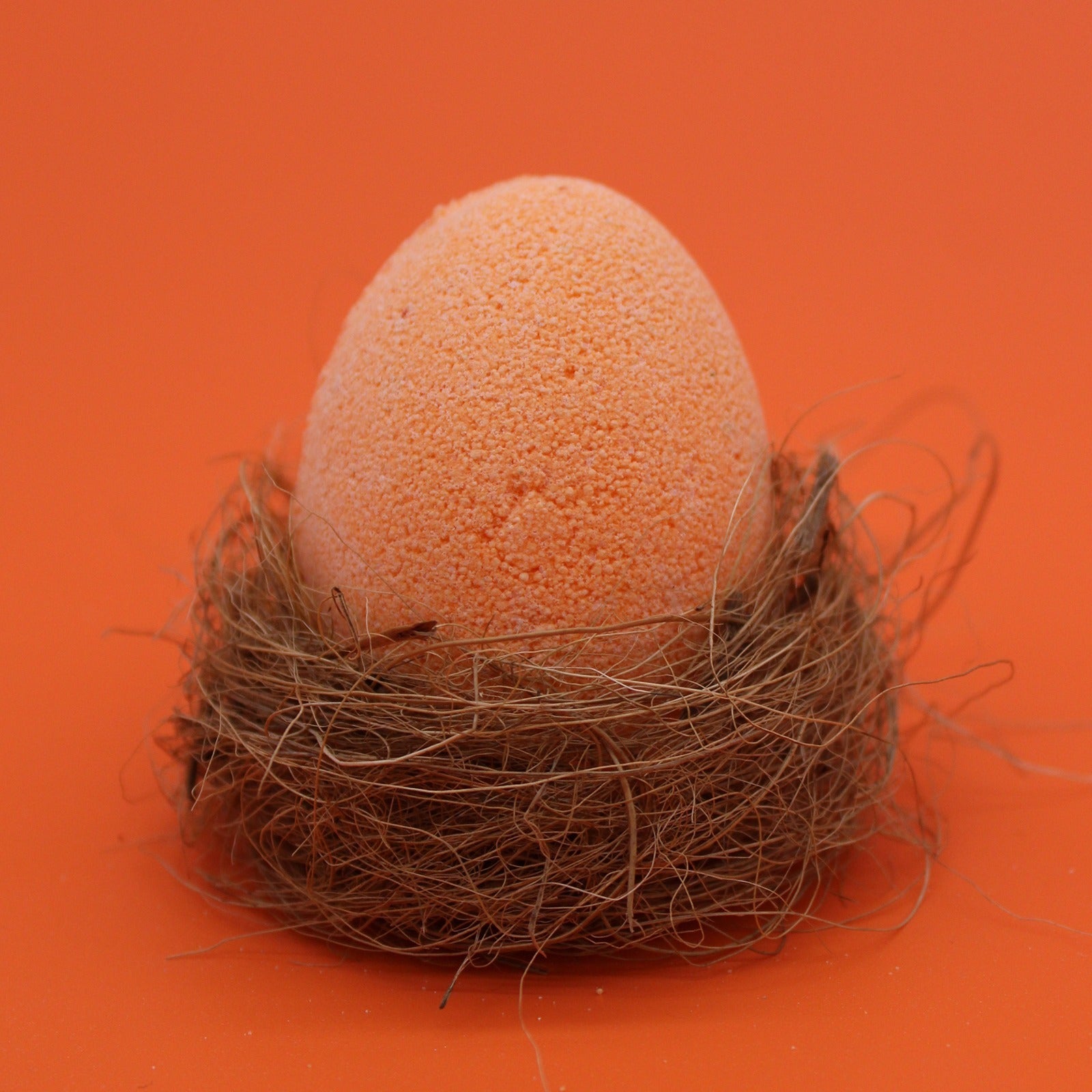 A textured orange egg-shaped object sits in a nest made of coarse, tangled fibers against a solid orange background.