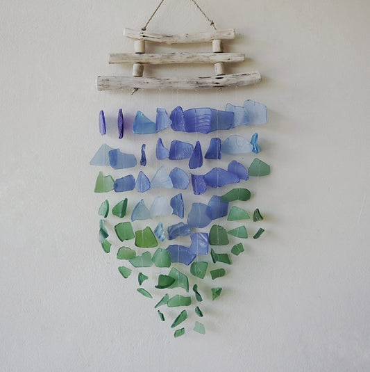 A sea glass wind chime hangs against a white wall. Numerous pieces of sea glass in shades of purple, blue, and green are suspended from a driftwood frame.