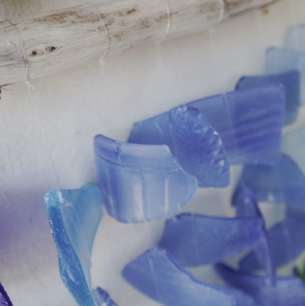 Sea glass pieces hang from a driftwood rod. They are various shades of blue and are suspended against a white background.