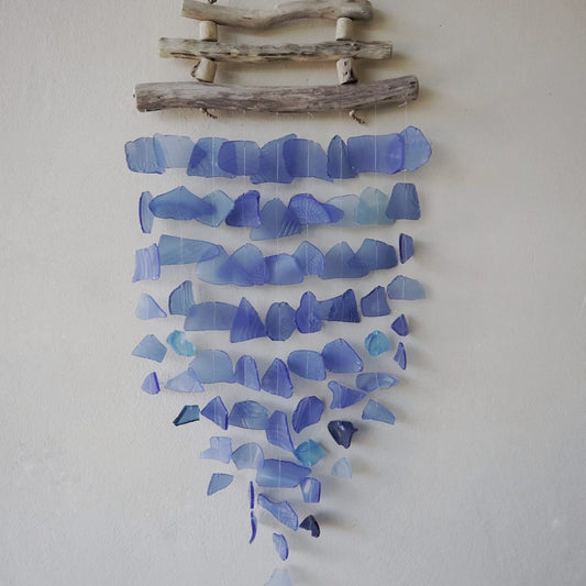 A sea glass mobile hangs from driftwood against a light wall. Numerous shades of blue glass pieces are strung in cascading rows.