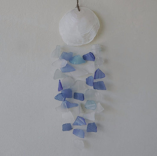 A seashell-topped mobile hangs against a wall. Many small, translucent blue and white glass pieces dangle below.
