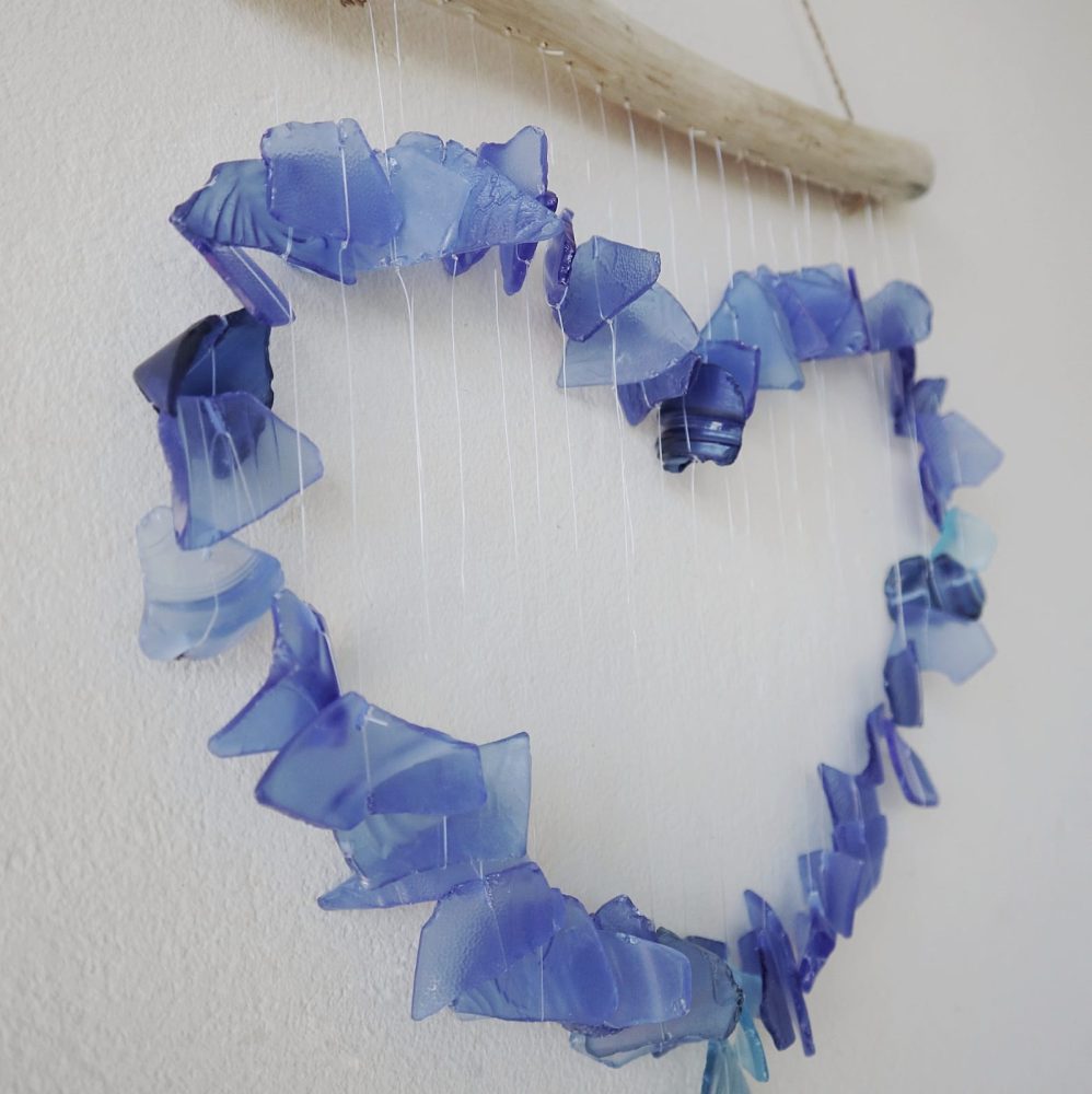 A heart-shaped mobile hangs on a wall. It is constructed from numerous pieces of translucent blue sea glass suspended from a driftwood branch.